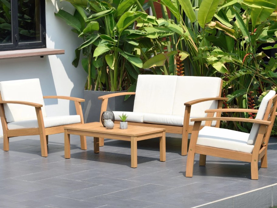 outdoor patio set in light wood with cream cushions, loveseat, 2 chairs and coffee table with plants behind them