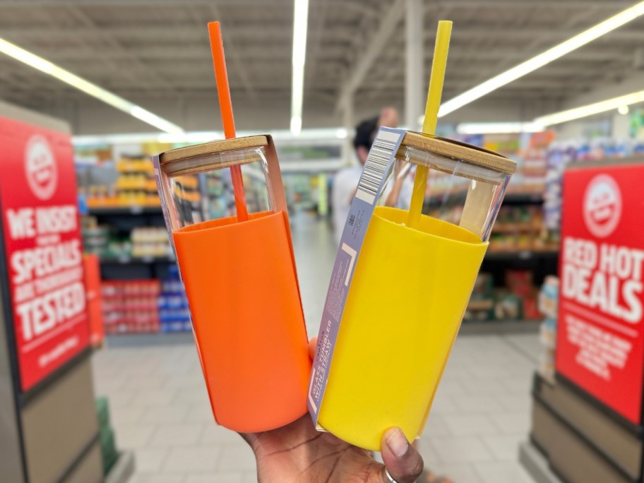 hand holding 2 glass tumblers with silicone sleeves and straws in orange and yellow