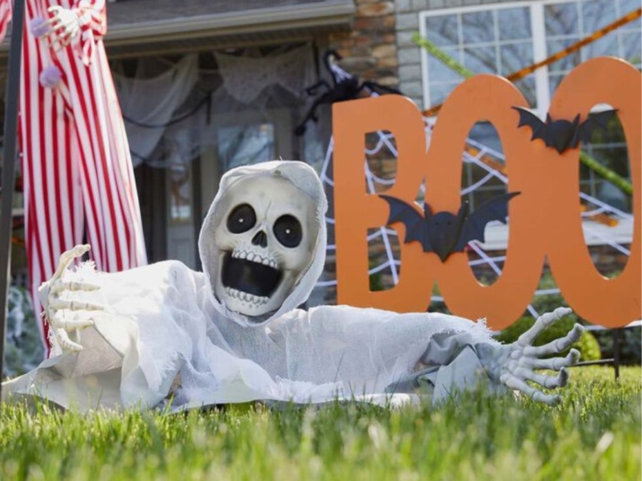 a yard with Halloween decor including a skeleton head, shoulders and arms coming out of the ground and a large orange sign that says "BOO" with black bats on it