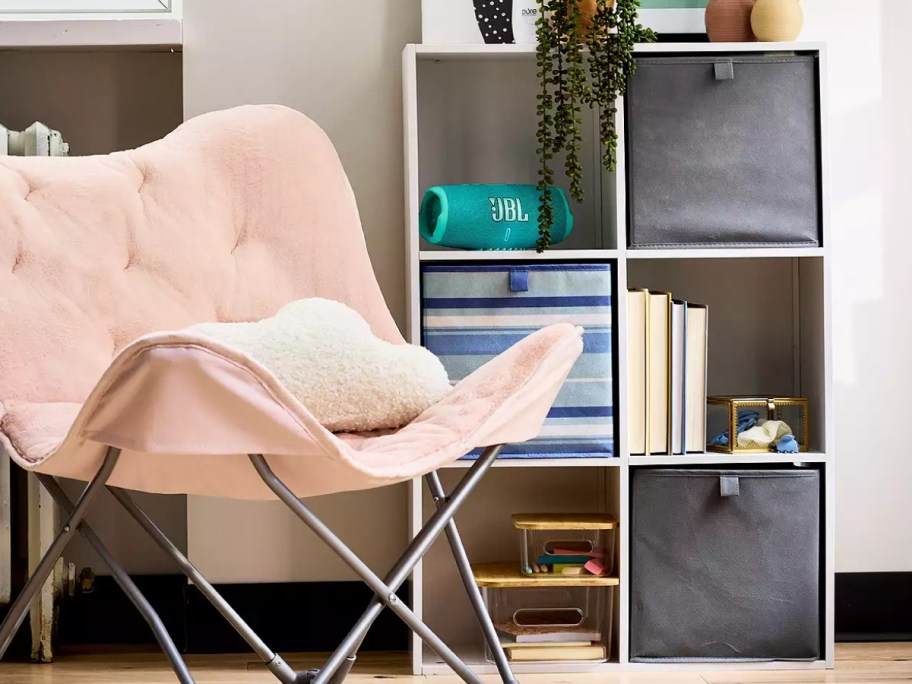 pink butterfly chair next to a white 6 cube organizer with grey and blue stripe organizer bins, books, and other items in it