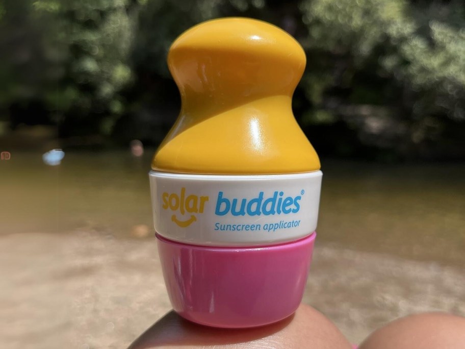 pink and yellow Solar Buddies sunscreen applicator on a person's knee, lake in the background