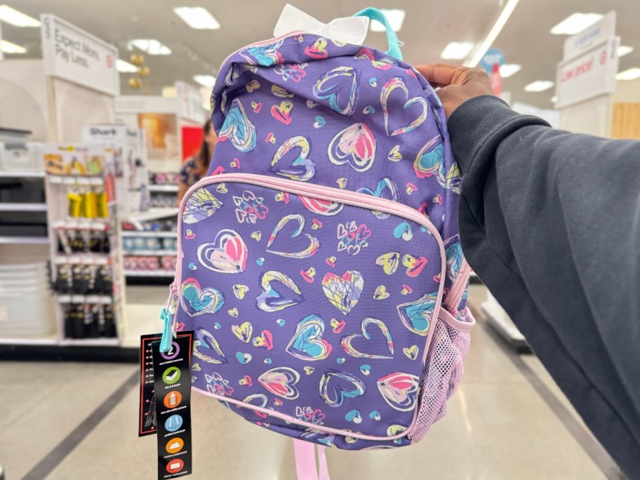 hand holding a kid's backpack that's purple with scribbled hearts in other colors