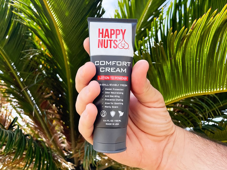 hand holding up bottle of Happy Nuts Comfort Cream in front of palm tree