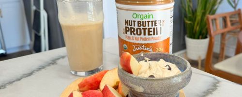 Orgain x Justin's Nut Butter Protein Powder next to a glass of protein shake and a cutting board with apple slices and a peanut butter dip