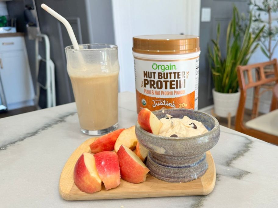 Orgain x Justin's Nut Butter Protein Powder next to a glass of protein shake and a cutting board with apple slices and a peanut butter dip