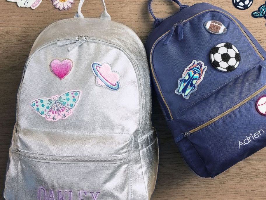 A Pottery Barn Kids Colby Silver Backpack with patches on it next to a smaller navy blue backpack with patches on it