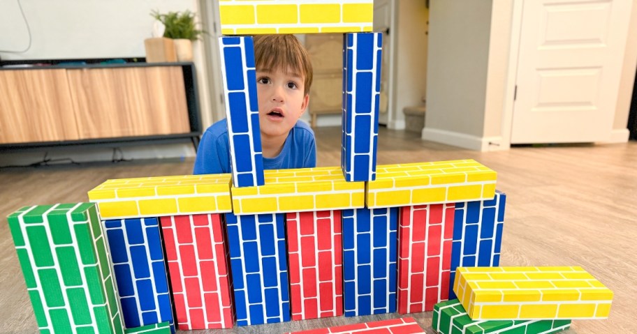 little boy peeking thru a tower built with primary color cardboard building blocks