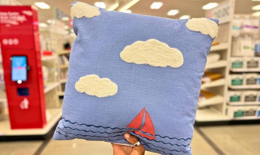 a womans hand holding a square throw pillow with a sail boat on it