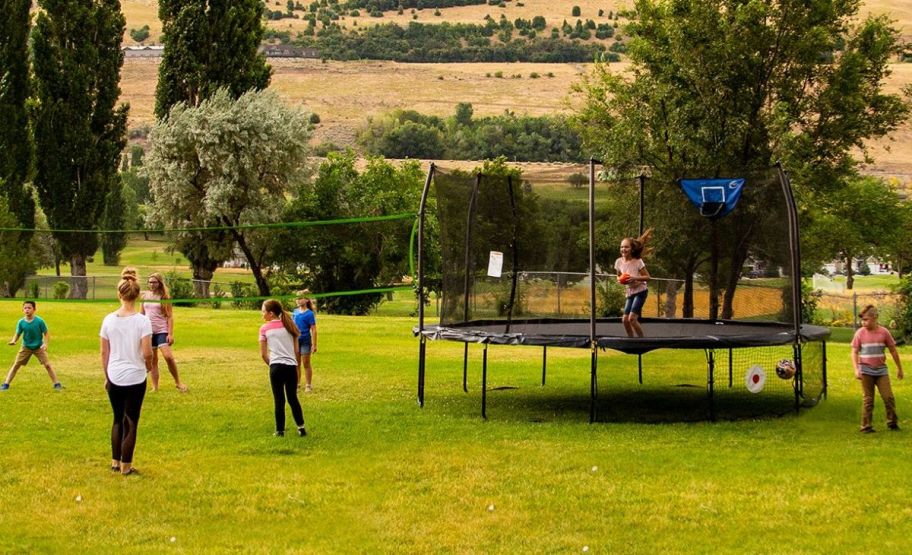 several kids playing on an enclosed trampoline with attached volley ball net in a back yard 