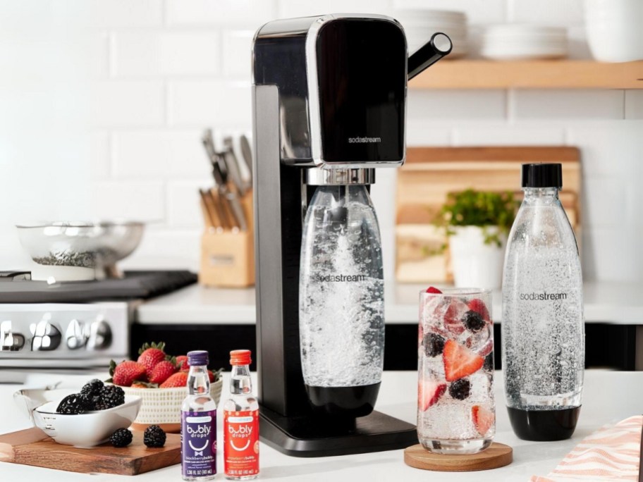 SodaStream Art Sparkling Water Maker with 2 reusable bottles, 2 Bubly flavor bottles and a glass of sparkling water, and bowls of fruit on a kitchen counter