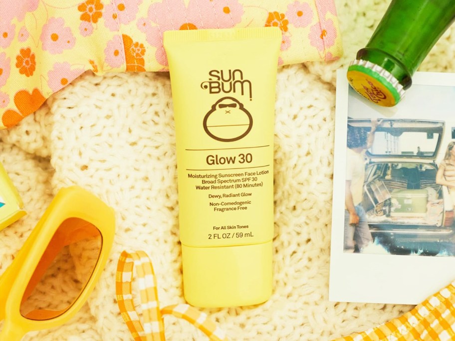 yellow bottle of sun bum glow face sunscreen on blanket near sunglasses and bathing suit