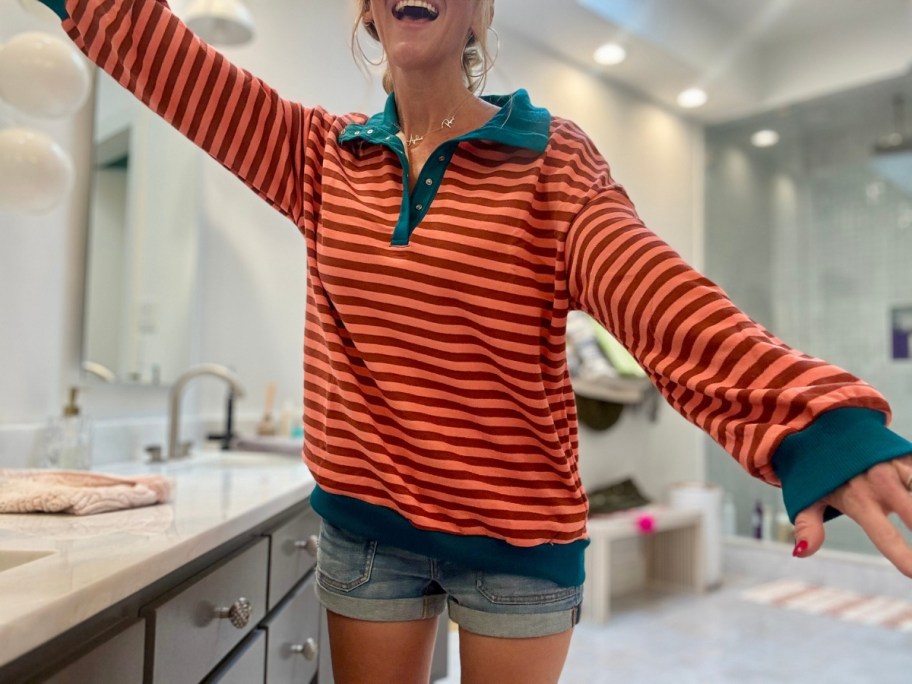 woman wearing a coral and orange striped sweatshirt that has a green collar and band around the sleeves and bottom with jean shorts