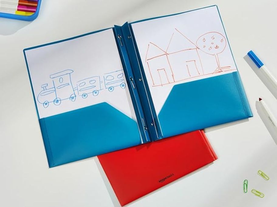 Amazon Basics Heavy-Duty 2-Pocket Plastic Folder 2-Pack on table with drawing in them and markers and paperclips around them