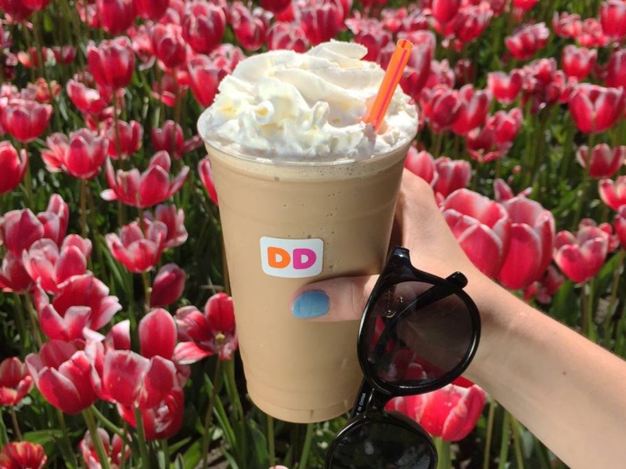 person holding Dunkin frozen coffee in hand with flowers in background