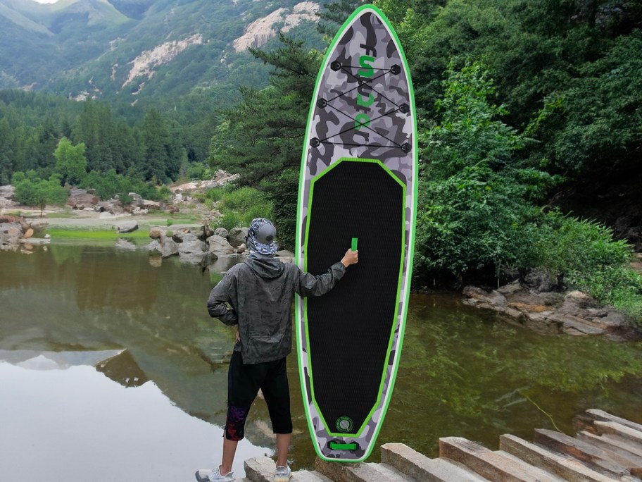 man holding up inflatable paddleboard in front of lake