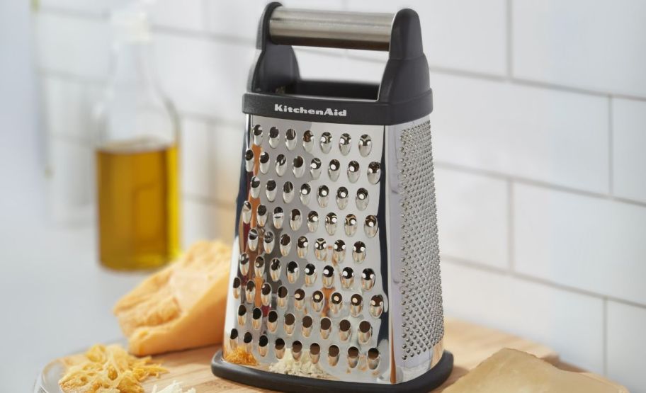 a steel box grater next to a cheese wedge and a bottle of olive oil