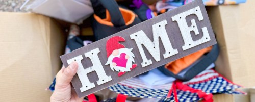 large box filled with various home goods, decor, tote bags, wrapping paper and hand holding a small wooden sign that says "Home" with a gnome on it
