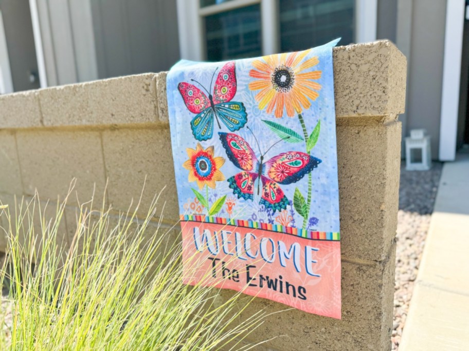personalized garden flag with butterflies and flowers hanging on a stone fence