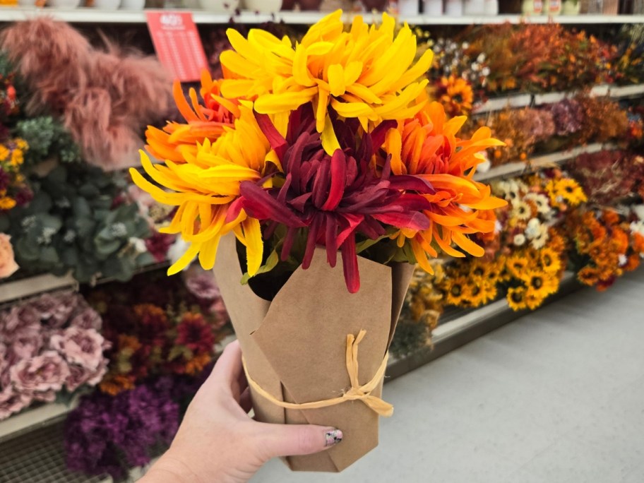 hand holding a faux mum plant in mixed fall colors or red, orange and yellow wrapped in brown kraft paper