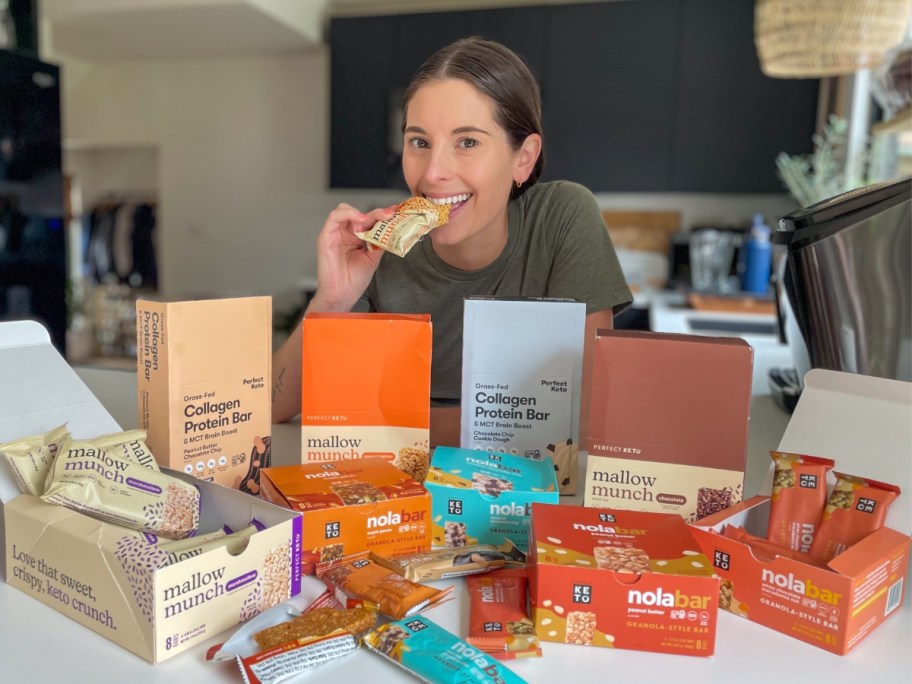 Emily taking a bite out of a Perfect Keto bar, with boxes and bars on a kitchen counter in front of her