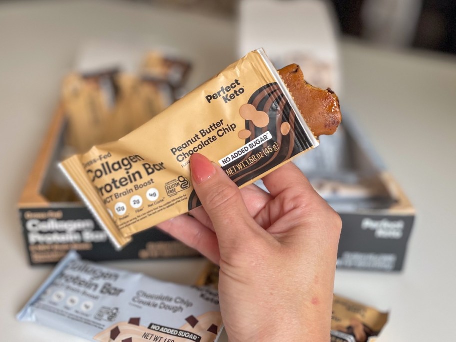 hand holding a Perfect Keto Collagen bar with a bite taken out of it, boxes of more bars behind it