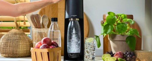 black SodaStream Terra on a kitchen counter surrounded by fruits, a glass of of sparkling water, an extra SodaStream bottle