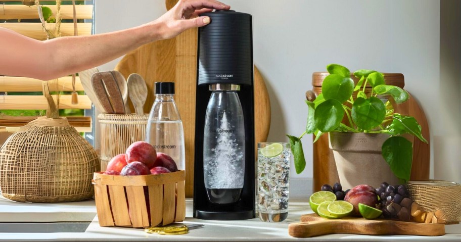 black SodaStream Terra on a kitchen counter surrounded by fruits, a glass of of sparkling water, an extra SodaStream bottle