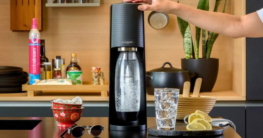 black SodaStream Terra Sparkling Water Maker in a kitchen, person's hand on it making a beverage, glass of ice beside it