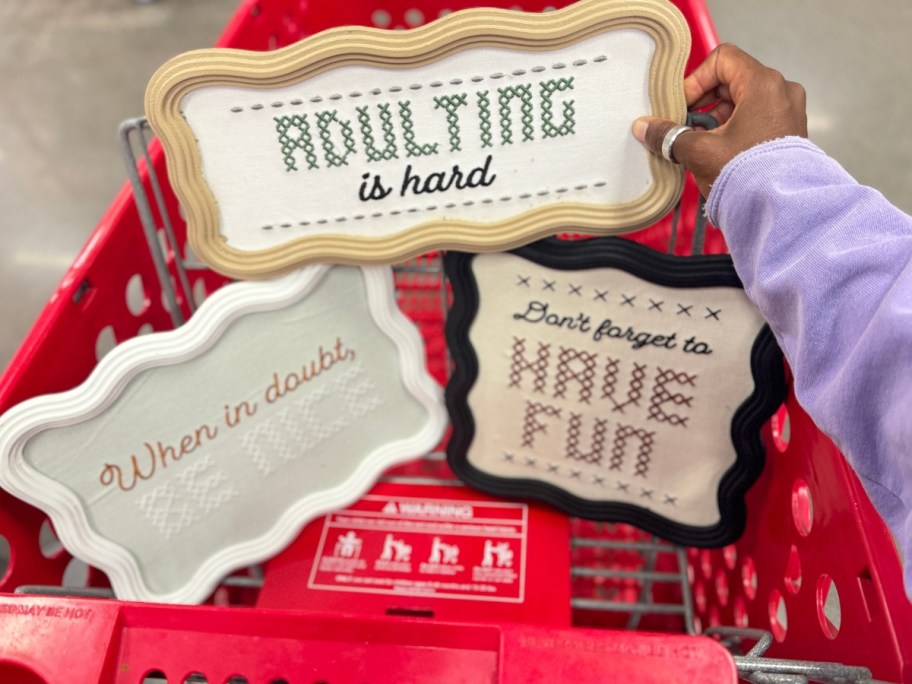hand reaching for wood wall signs with funny phrases in cross-stitch that are sitting in a Target cart