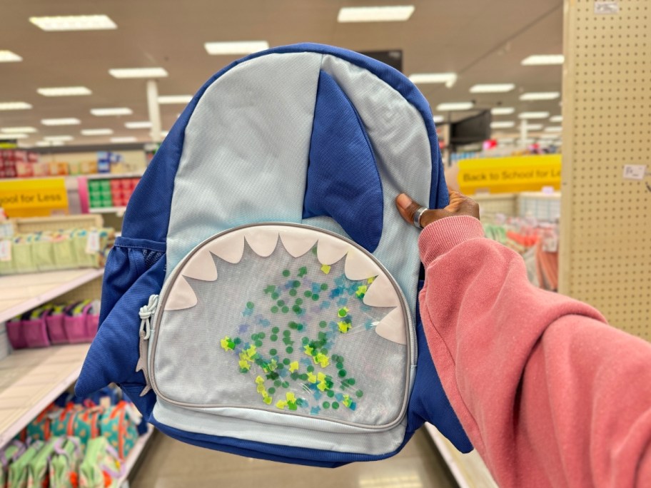 hand holding a kid's blue backpack that looks like a shark with fins and teeth