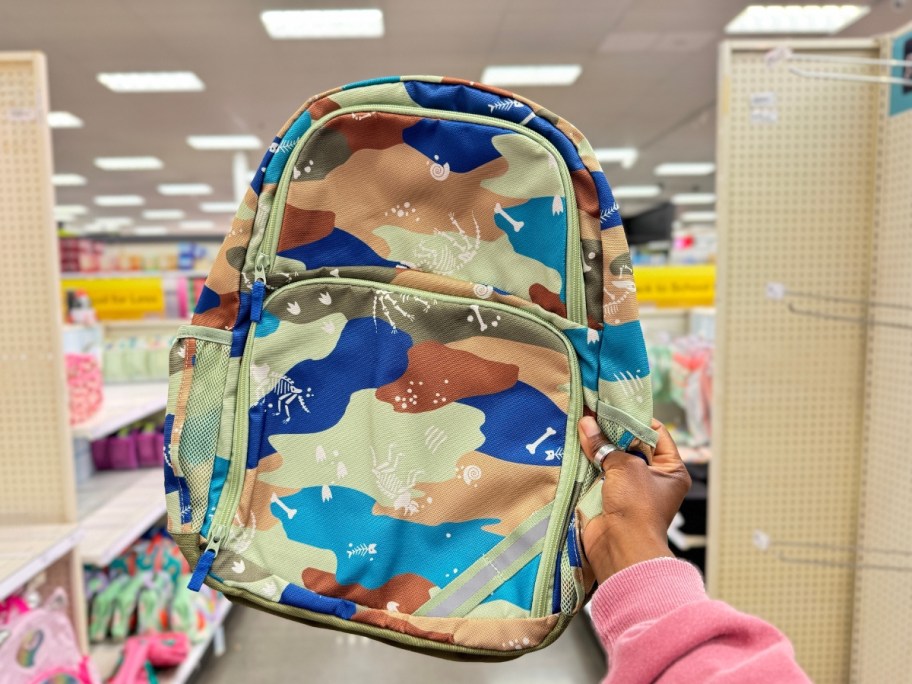 hand holding a kid's backpack in greens, blues and browns with Dinosaurs on it