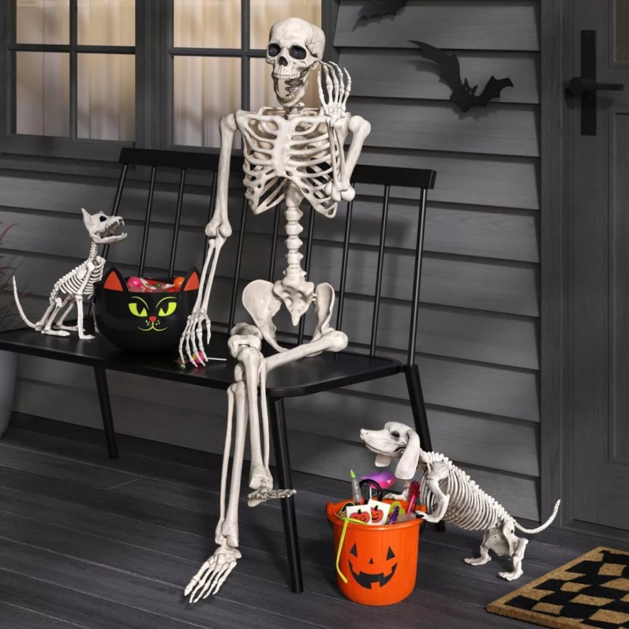6 foot posable white skeleton Halloween decoration sitting on a bench on a front porch, skeleton dogs beside him