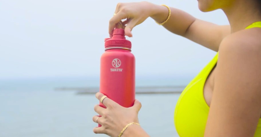 Woman standing by the ocean wearing a yellow sports bra and holding a bright pink water bottle