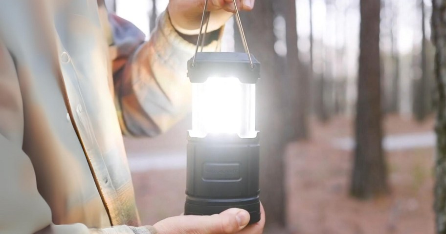 man wearing a flannel shirt holding a camping lantern that's lit up standing in the woods