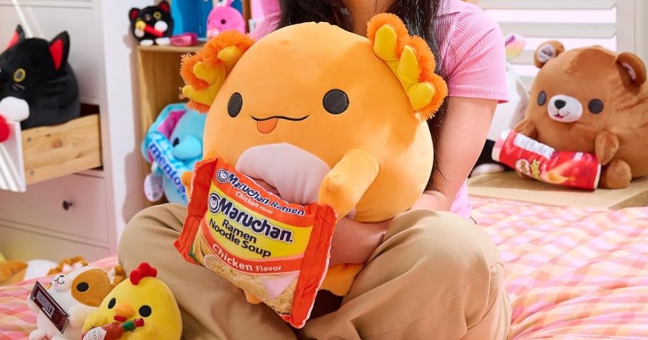 girl sitting on a bed holding a large Snackles axolotl plush, with more Snackles around her