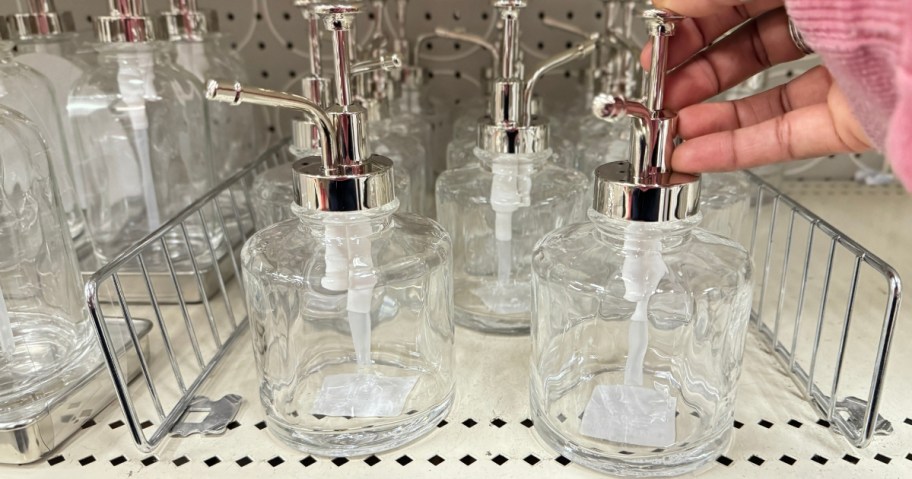 hand reaching for a clear glass and silver oil spout pump style soap dispenser on a store shelf