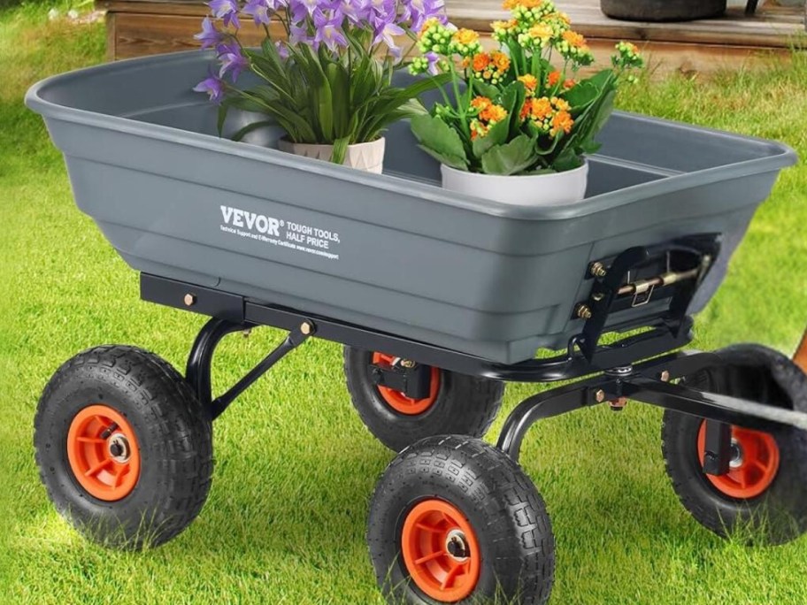 grey garden dump cart with black and orange wheels filled with flowers in plastic pots