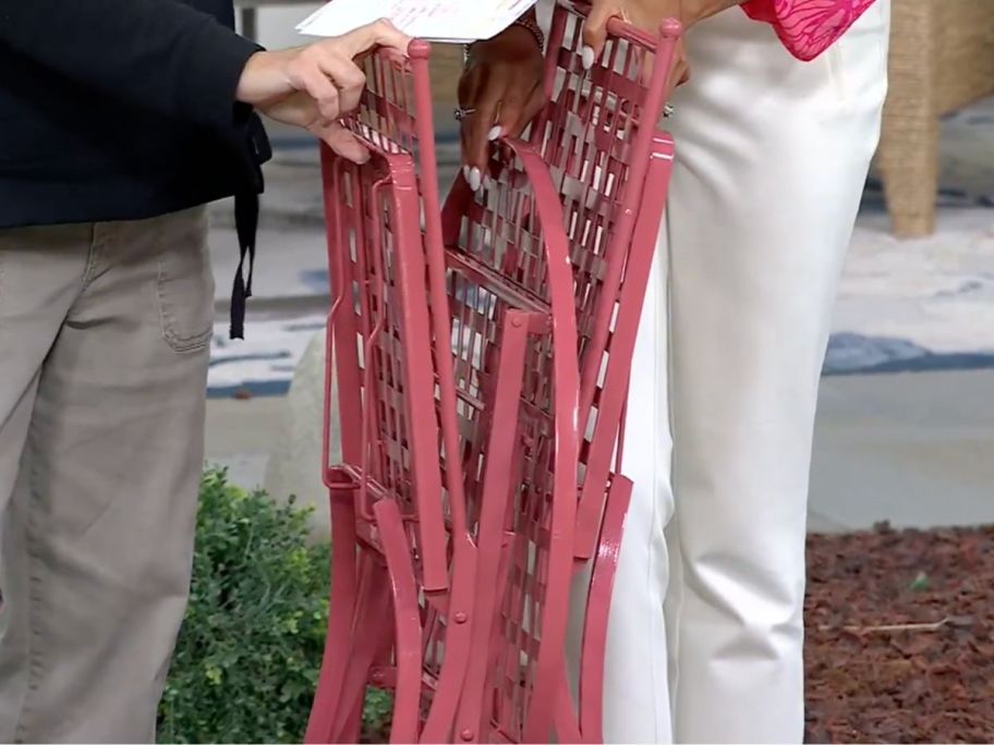 A foldable metal bistro set with all 3 pieces folded flat held up between two women