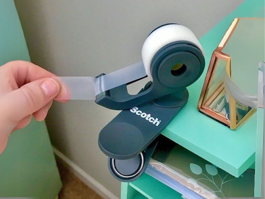 hand pulling tape from Scotch Tape Dispenser attached to desk