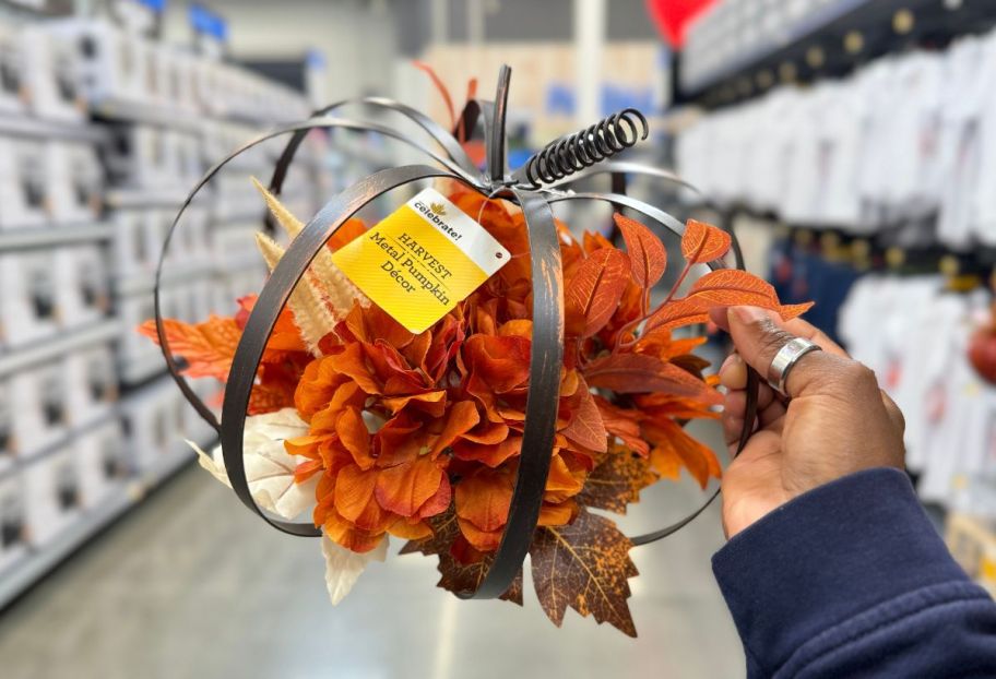 a womans hand holding a metal framed pumpkin shaped fall floral decor