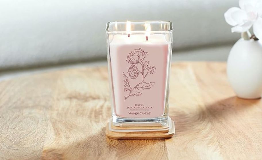 a large pink candle in a square glass jar sitting on a table next to a flower vase.