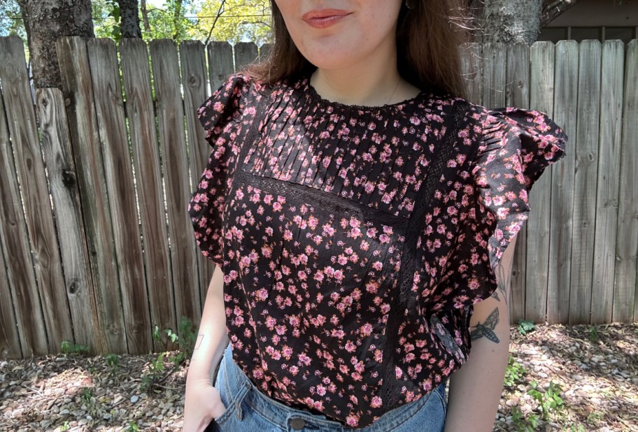 woman wearing a flutter sleeve top and jeans, standing in front of a fence