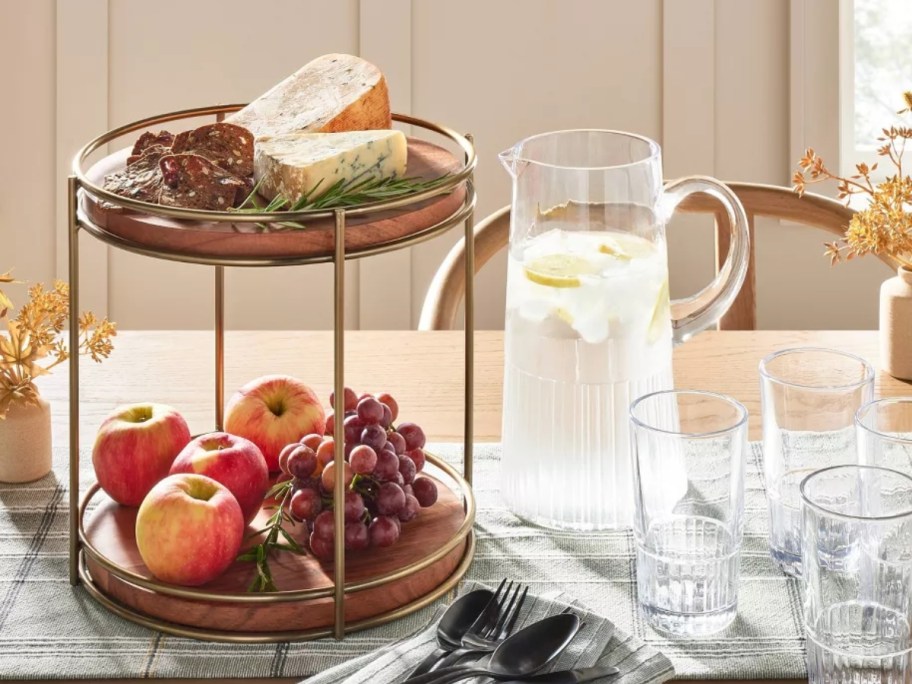 2-tiered wood serving tray with brass accents filled with fruit and cheeses next to a glass pitcher of water on a table