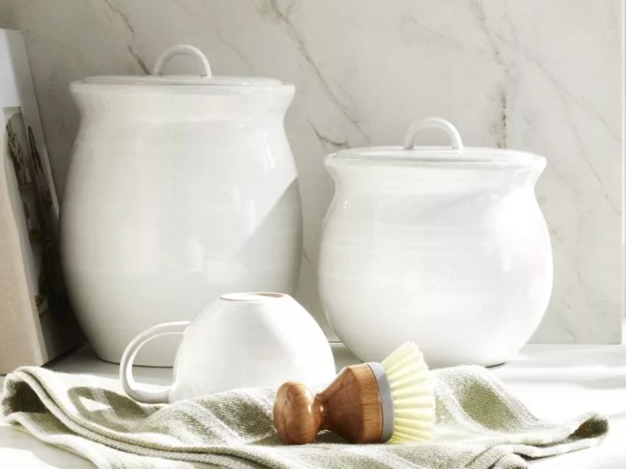 medium and large cream color vintage style kitchen canisters on a counter, upside down coffee mug, dish scrubber brush and dish towel in front of them