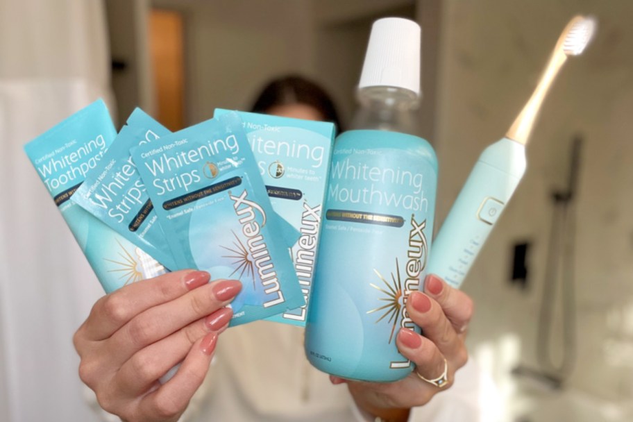 woman holding mouthwash, teeth strips, toothpaste, and toothbrush in front of face