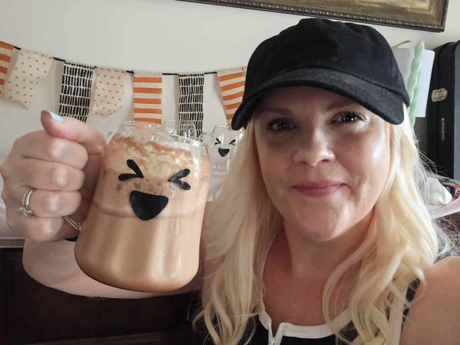 smiling woman holding up a glass ghost mug filled with hot chocolate in front of a fall bunting banner