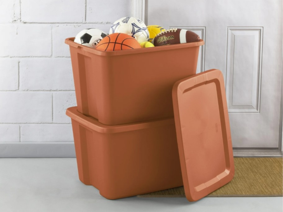 Two large brown plastic storage totes stacked on top of each other, one with a lid on the side and filled with sports balls.
