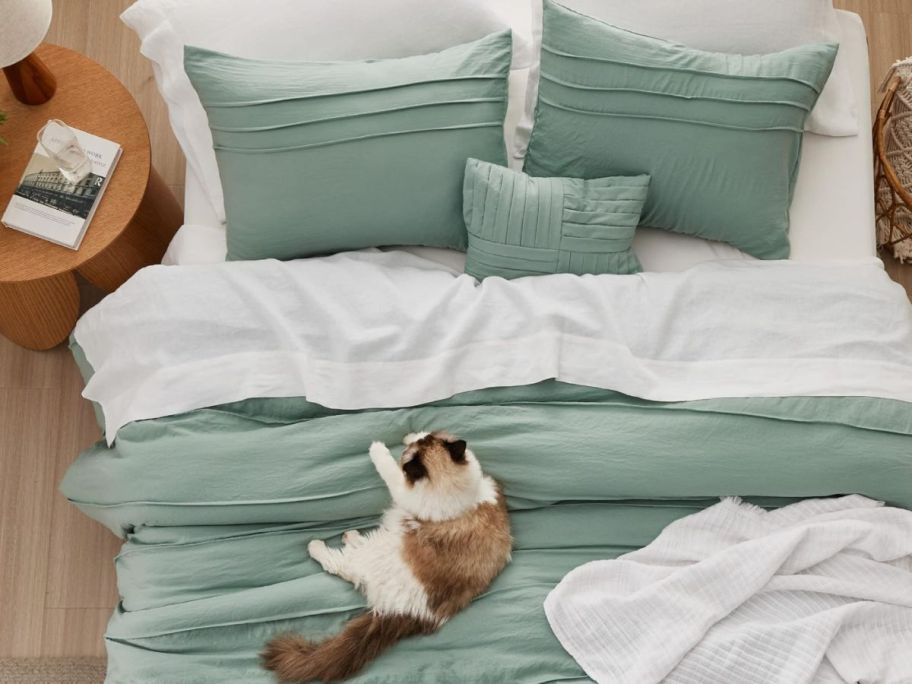 A cat waking up in a comfy bed with green pillows and a comforter