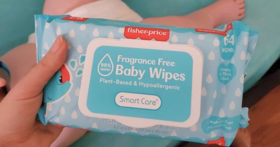 hand holding a blue pack of baby wipes, baby laying in crib behind it