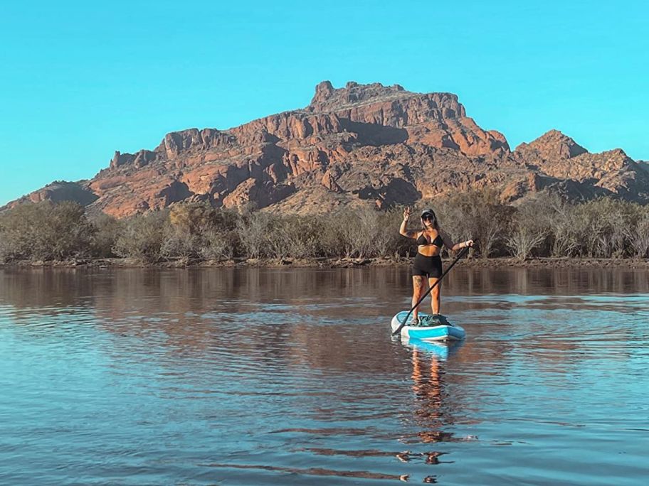 Woman standing on a Funwater Paddle Board in water while giving a peace sign with a mountain in the background